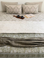 A styled bed dressed in neutral-toned bedding, featuring a cream quilted top layer, an olive green patterned lower sheet, matching patterned pillows, and a textured olive throw blanket draped across the foot of the bed. A small wooden tray with two dark cups and an open book rest on the bed.