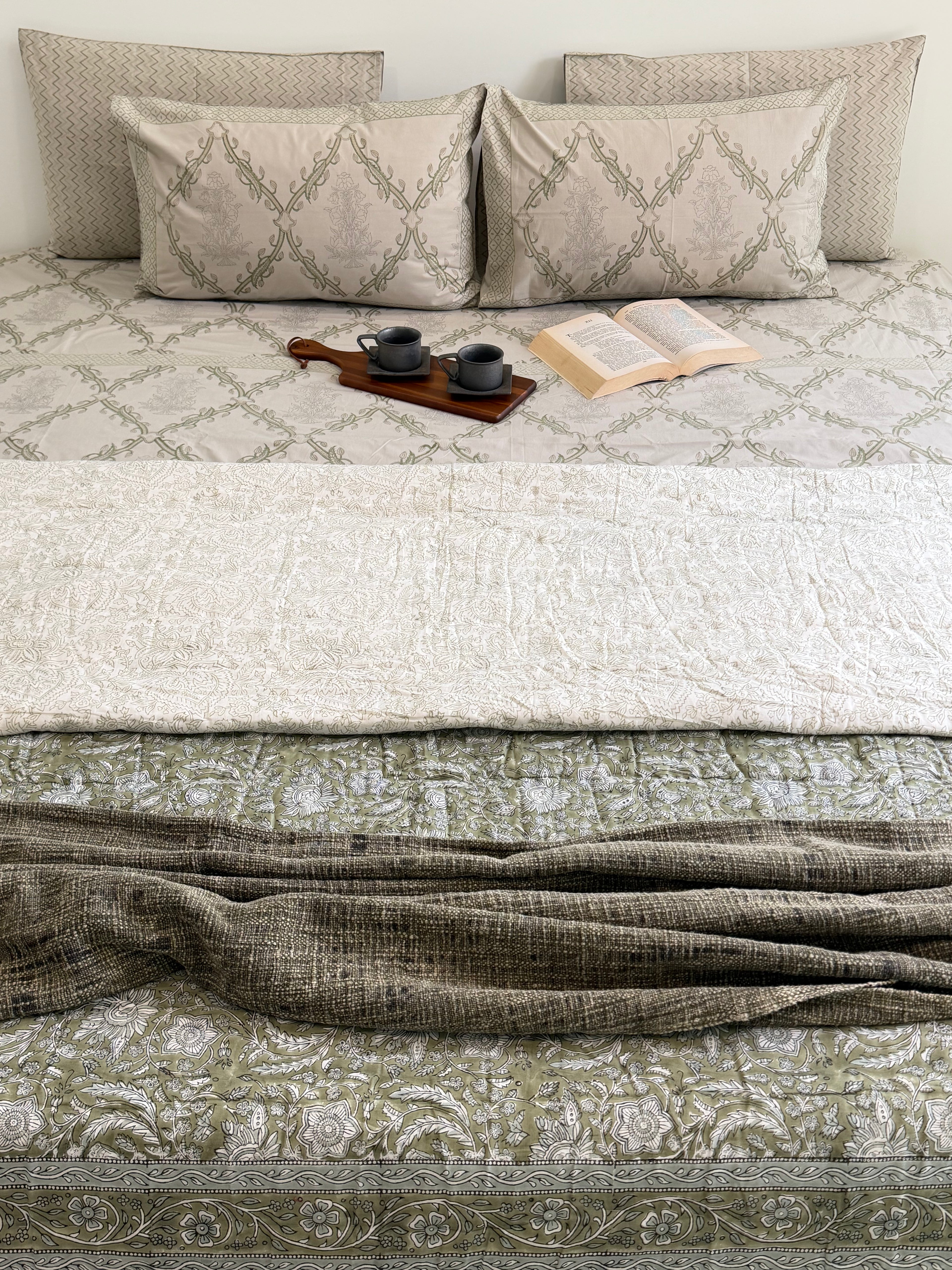 A styled bed dressed in neutral-toned bedding, featuring a cream quilted top layer, an olive green patterned lower sheet, matching patterned pillows, and a textured olive throw blanket draped across the foot of the bed. A small wooden tray with two dark cups and an open book rest on the bed.
