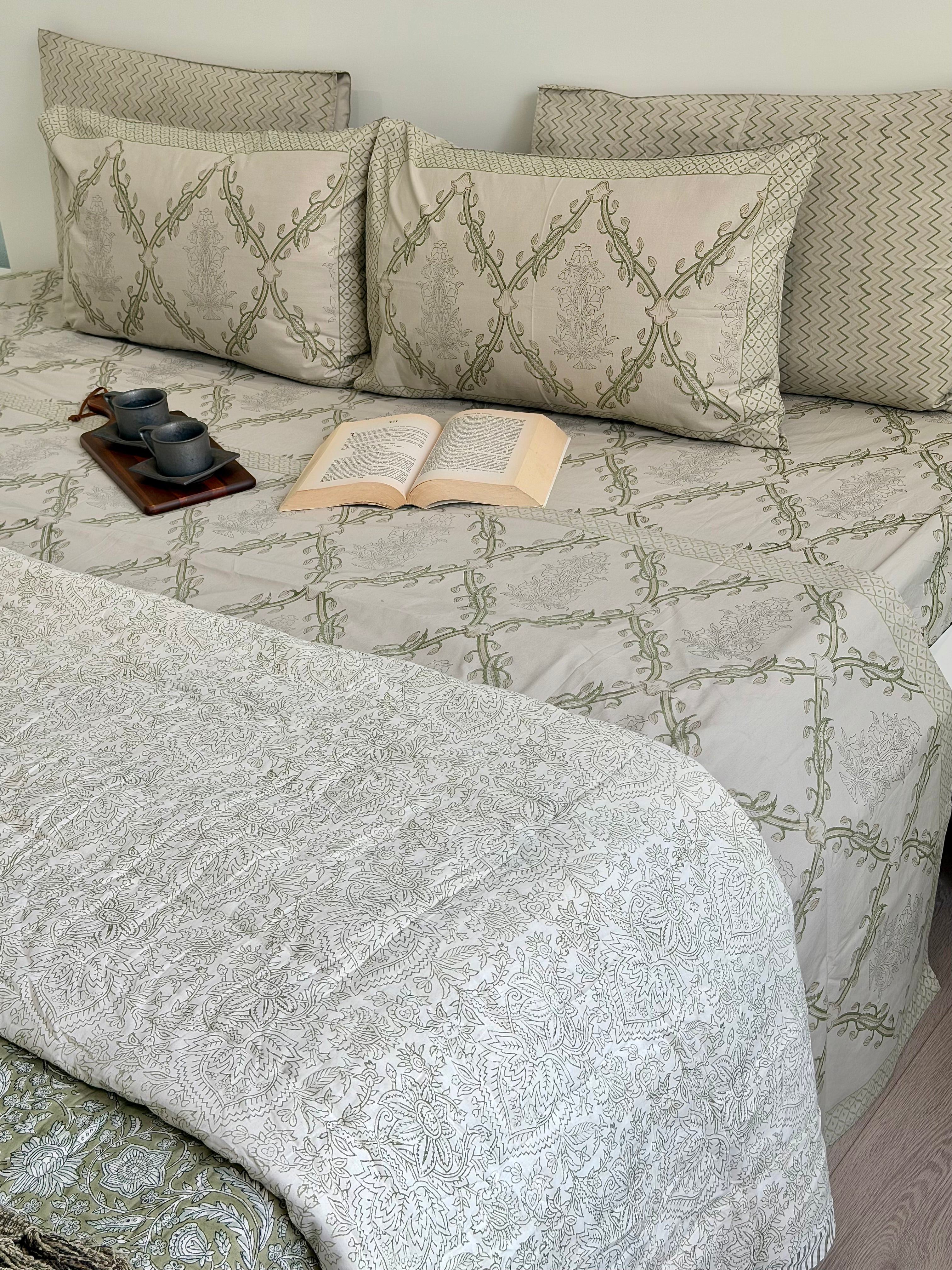 A neatly made bed with a light beige and sage green damask-patterned quilt, matching pillow shams, and solid patterned pillows. An open book and a small cup and saucer set are styled on the bed.